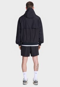 Czarna, lekka kurtka z kapturem, elastycznymi mankietami i pasem, zestawiona z czarnymi szortami. Posiada delikatny połysk i luźny krój.