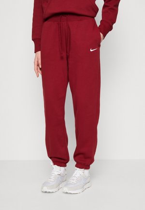 Osoba ubrana w bordowe spodnie dresowe Nike ze ściągaczem w talii i białe buty sportowe Nike, stojąca na jednolitym jasnym tle.