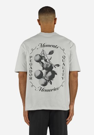Άνδρας που φοράει λευκό μπλουζάκι με μαύρο σχέδιο κλαδιού φρούτων και κείμενο «Moments», «Pegador», «Quality» και «Memories» στην πλάτη.