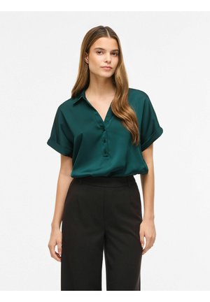Femme aux longs cheveux bruns portant un chemisier vert foncé à manches courtes et un pantalon noir taille haute sur un fond blanc uni.