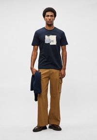 T-shirt de gola redonda azul-marinho com um design impresso em cores claras. Combinado com calças largas em tom de bege e segurando um moletom azul-marinho. Sapatos castanhos.