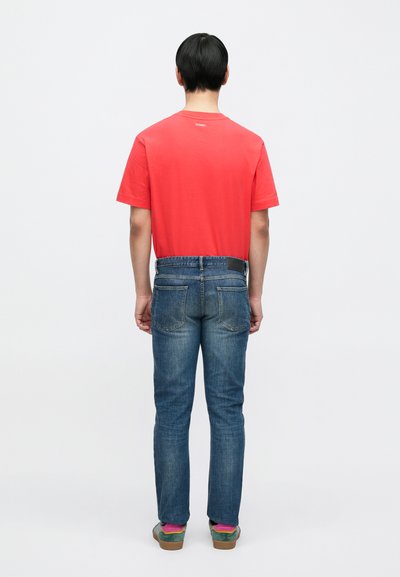 T-shirt en coton rouge, coupe droite, associé à un jean en denim bleu avec deux poches arrière et un patch en cuir noir à la taille.