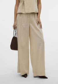 Vrouw draagt beige wijde broek en bijpassend topje, met een kleine donkerbruine handtas in de hand, staande tegen een effen achtergrond.