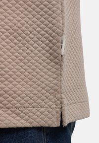 Chaqueta de tela acolchada beige con abertura lateral y pequeña etiqueta blanca que dice "EST. 1903", llevada sobre vaqueros de mezclilla azul oscuro.