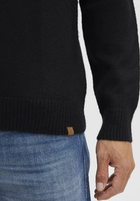 Schwarzer, gestrickter Pullover mit gerippten Bündchen und Saum. Verfügt über ein kleines braunes Markenpatch nahe dem unteren Ende. Getragen über blauen Jeans.