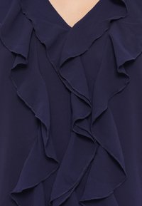 Blusa blu scuro con volant sovrapposti lungo il colletto, realizzata in un tessuto leggero e liscio con una finitura morbida.