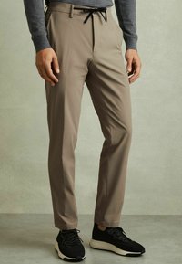Pantaloni bej conici cu un sistem de prindere cu siret, textură netedă și buzunare laterale, asortați cu pantofi sport negri.