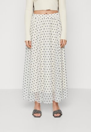 Kvinde iført en hvid ankellang nederdel med sorte polkaprikker, en cremefarvet ribstrikket langærmet croptop og sorte slidesandaler med spænder.