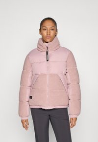 Woman standing wearing a pink quilted and ribbed high-neck jacket and dark pants against a plain light background.