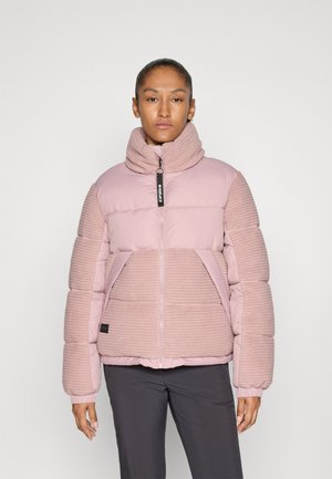 Kvinne som står iført en rosa quiltet og ribbet høyhalset jakke og mørke bukser mot en ensfarget lys bakgrunn.