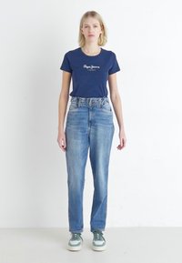 Marineblauwe t-shirt met ronde hals en wit logo, hoog opgesneden lichtblauwe jeans en groen-witte sneakers. Eenvoudig ontwerp, casual pasvorm.