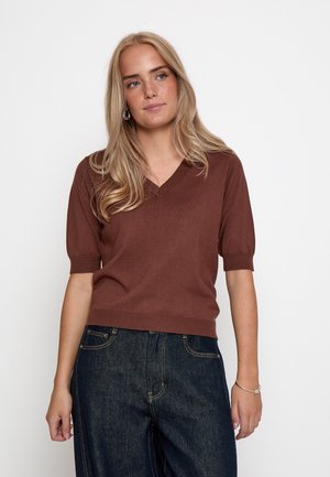 Blonde vrouw met een bruine V-hals trui met korte mouwen en donkerblauwe spijkerbroek, staand tegen een witte achtergrond.