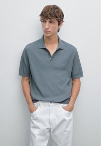 Szara koszulka polo z krótkim rękawem o fakturowanym, prążkowanym wzorze, zapinana na trzy guziki, o luźnym kroju, zestawiona z białymi spodniami.