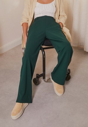 Person iført mørkegrønne vidde bukser, beige slip-on sko, hvid top og beige cardigan, der sidder på en sort stol med hjul indendørs.