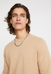 Beige neule, jossa on teksturoitu neulos, pyöreä pääntie ja ribbaatut hihansuut. Mukana tummasta ketjusta valmistettu korvakoru. Mallilla on kihara hius ja neutraali ilme.