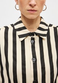 Πουκάμισο με κουμπιά, με κάθετες μαύρες και κρεμ ρίγες, κλασικό γιακά και μαύρα κουμπιά στο μπροστινό μέρος. Λείο ύφασμα.