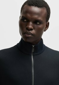 Close-up of a dark navy zipped jacket with a high collar, made from textured fabric, featuring a silver zipper and minimalistic design elements.