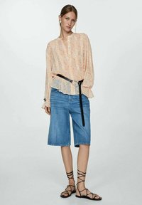 Bloemige, langemouwen, lichtoranje blouse met knoopsluiting, gecombineerd met blauwe denim short en zwarte sandaaltjes met banden.