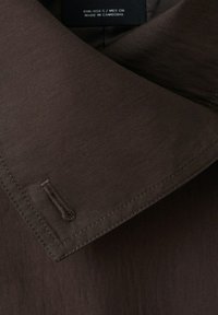 Cuello de abrigo marrón oscuro con costuras visibles de ojal y una etiqueta negra que muestra la talla y el texto "Hecho en Camboya".