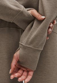 Top din tricot cu model în structură de vafă, cu mâneci lungi, de culoare taupe, care pune în evidență un design texturat. Manșetele au deschideri pentru degete, iar mâinile sunt parțial vizibile.