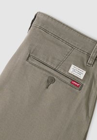 Hlače znamke Levi's v kaki barvi, prikaz zadnjega žepa z gumbno zaponko in dvema etikama znamke Levi's, ena bela in ena rdeča.