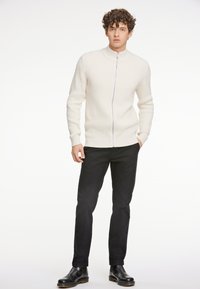 Krämfärgad stickad jacka med ståkrage och framdraget blixtlås, kombinerad med mörka byxor och svarta ankelstövlar. Modellen står i en neutral miljö.