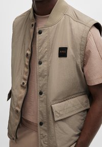 Chaleco sin mangas de color beige claro con un acabado texturizado, cierre de cremallera y automáticos, y un bolsillo frontal. Incluye un parche de marca negro en el pecho.