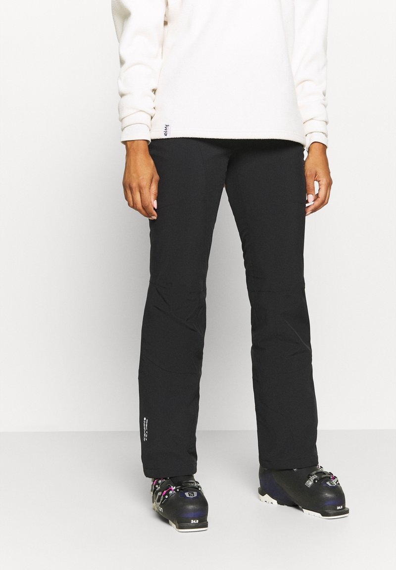Personne portant un pantalon de ski noir et des bottes de ski noires avec des boucles roses et argentées, debout sur un fond blanc.