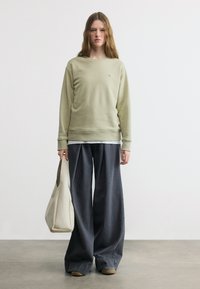 Νεαρό άτομο με μακριά μαλλιά, φοράει ανοιχτοπράσινο φούτερ, φαρδιά σκούρα γκρι παντελόνια, κρατώντας μια μεγάλη εκρού τσάντα, στέκεται μπροστά σε ένα απλό φόντο.