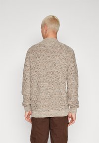 Rudas megztinis su tekstūruotu deimanto raštu, su banguotais rankogaliais ir apačia, turintis apvalią iškirptę. Dėvimas su rudomis kelnėmis.