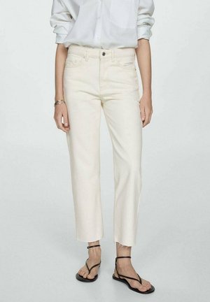 Vrouw draagt high-waist crèmekleurige jeans met onafgewerkte zomen, wit hemd met lange mouwen, gouden armbanden en zwarte sandalen met bandjes.