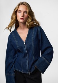 Denimjakk on V-kaelusega, ees tõmblukuga ja laiade varrukatega, millel on nähtav õmblus. Sügav sinine värv, sile tekstuur, vabaaja stiil.