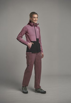 Woman wearing a mauve and black zip-up jacket with hood, mauve pants, and hiking shoes, standing with one hand in pocket against plain background.