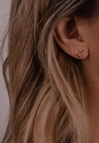 Na uhu su vidljive naušnice u obliku srca od ružičastog zlata, s glatkim, tankim žičanim dizajnom. Kosa je valovita i opušteno se spušta oko uha.