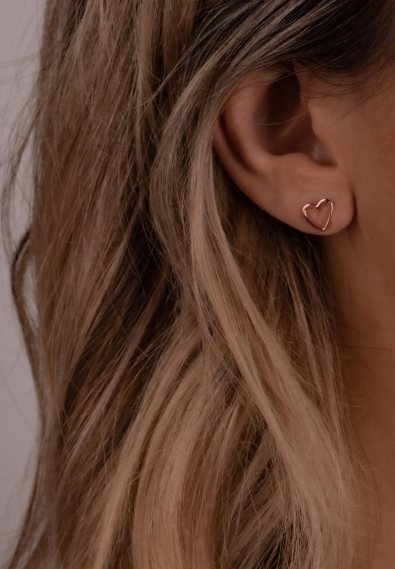 Na uhu su vidljive naušnice u obliku srca od ružičastog zlata, s glatkim, tankim žičanim dizajnom. Kosa je valovita i opušteno se spušta oko uha.