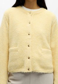 Cárdigan amarillo hecho de tela suave y texturizada, con escote redondeado, seis botones de tono dorado y dos bolsillos delanteros.