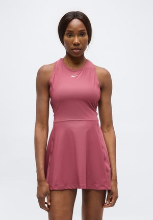 Femme athlétique à la peau et aux cheveux foncés portant une robe sans manches rose de Nike, face à l'avant sur un fond blanc uni.