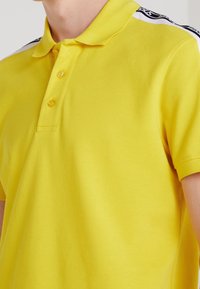 Polo jaune en tissu texturé, avec un col classique, trois boutons, et des accents rayés noir et blanc contrastants sur les épaules.