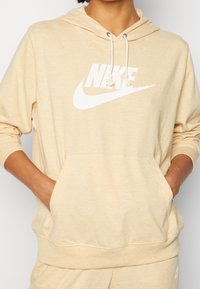 Světle béžová mikina s kapucí, přední kapsou a bílým logem Nike tištěným na hrudi. Měkká, směsová textura látky.