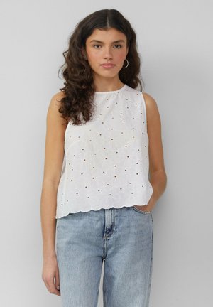 Weißes ärmelloses Oberteil mit floralem Durchbruchsdesign und gewelltem Saum. Getragen mit hellblauen Jeans, die einen lässigen Schnitt und strukturierte Stoffe aufweisen.