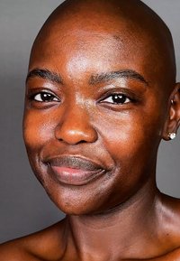 Skallig individ med slät, mörk hud, markerade kindben, bruna ögon och subtil läppform; bär små örhängen.