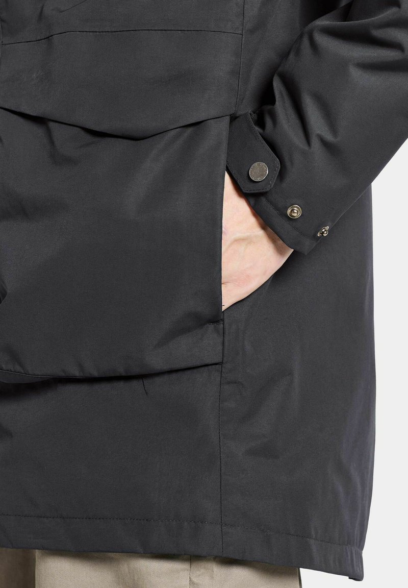 Schwarze wasserdichte Jacke mit einer Brusttasche, Druckknopfverschlüssen an den Manschetten und einer glatten Textur. Sichtbare Hand in der Seitentasche.