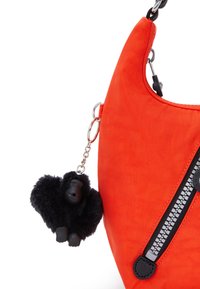Orange nylonhandväska med en böjd form, prydd med en svart dragkedja och en avtagbar svart gorillanyckelring som är fäst med en kedja.