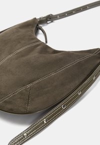 Suede handbag in dark olive green, crescent shape with white stitching. Features a long strap with metal accents and a zipper closure.