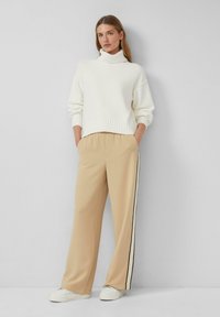 Cremfarbener Rollkragenpullover mit gerippten Bündchen, kombiniert mit beigen, weit geschnittenen Hosen mit seitlichen Streifen. Das Model trägt weiße Sneakers.