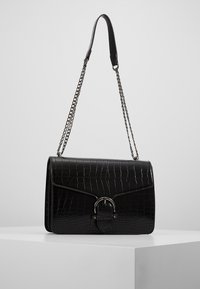 Schwarze rechteckige Handtasche mit glänzendem Krokodil-Präge-Design, silberner Kettenhenkel und einem runden Metallverschlussakzent auf der Vorderseite.