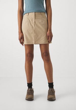 Persona che indossa una gonna beige al ginocchio, una camicia turchese, calzini neri e scarpe da trekking marroni, in piedi davanti a uno sfondo semplice.