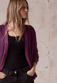 Violet ribbade koftor med knappstängning, bärs över en svart topp. Modellen poserar med händerna i fickorna mot en neutral bakgrund.