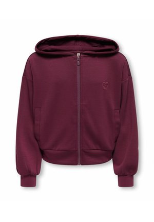 Fille aux longs cheveux bruns portant un sweat à capuche bordeaux zippé, un t-shirt blanc, un pantalon large noir et des bottes noires, debout avec une main sur la hanche.