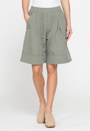 Donna che indossa pantaloncini verde oliva larghi con grandi risvolti, abbinati a scarpe beige senza lacci, in piedi con una mano in tasca.